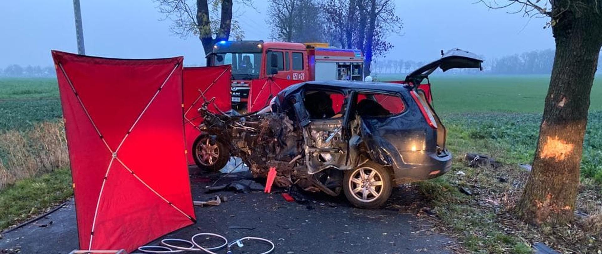 Zdjęcie przedstawia samochód osobowy ze zniszczonym lewym bokiem. Po lewej stronie auta znajduje się czerwony parawan, obok parawanu na niebieskiej macie znajdują się narzędzie hydrauliczne strażaków. Z tyłu stoi wóz strażacki.