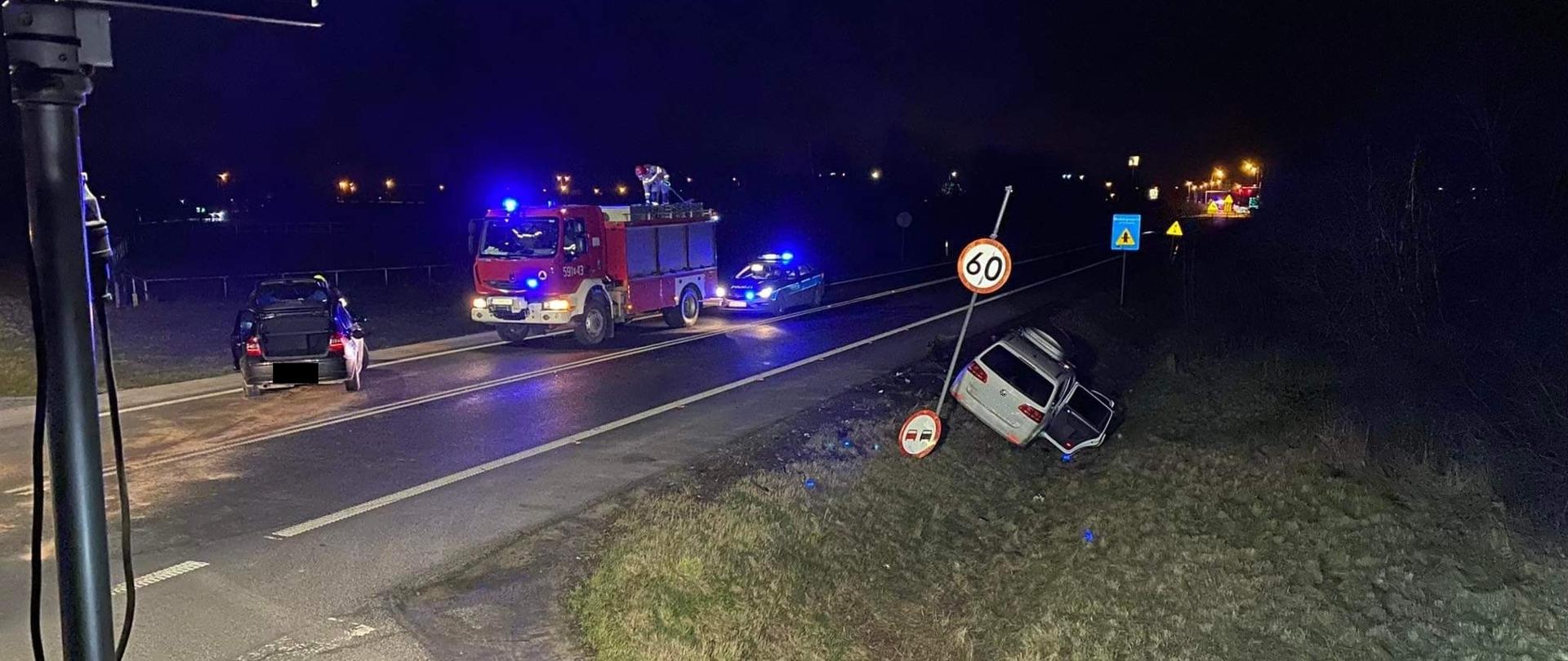 Widać dwa uszkodzone samochody, jeden w rowie, drugi na drodze, jest ciemno