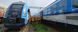 Strażacy stojący pomiędzy pociągami koordynują przejście pasażerów ze składu unieruchomionego do rezerwowego, w tle pociągi przewoźnika