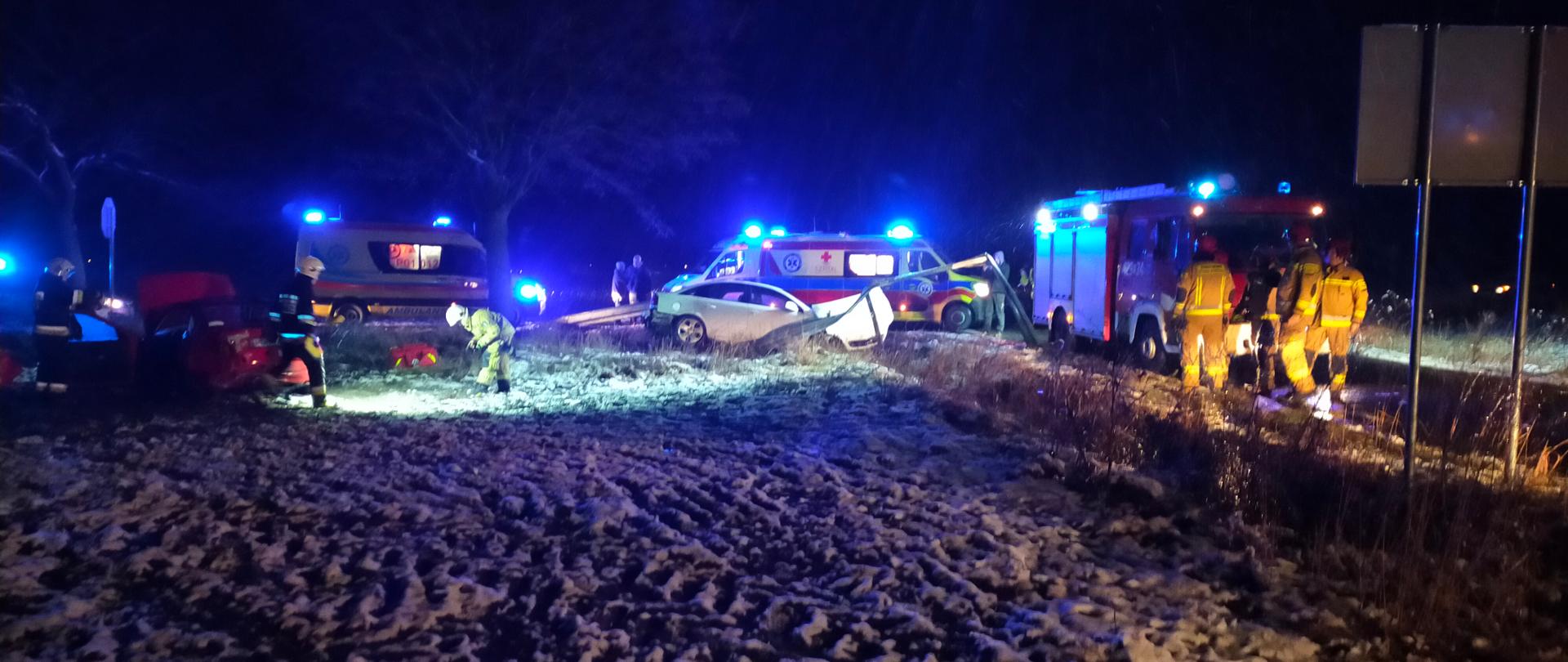 Na zdjęciu widać dwa samochody osobowe, które po zderzeniu zatrzymały się na przydrożnym polu w pozycji na kołach. Po prawej stronie widać samochód strażacki przed którym stoją strażacy a w tle dwie karetki pogotowia ratunkowego. Na polu widać lekki śnieg, jest ciemno