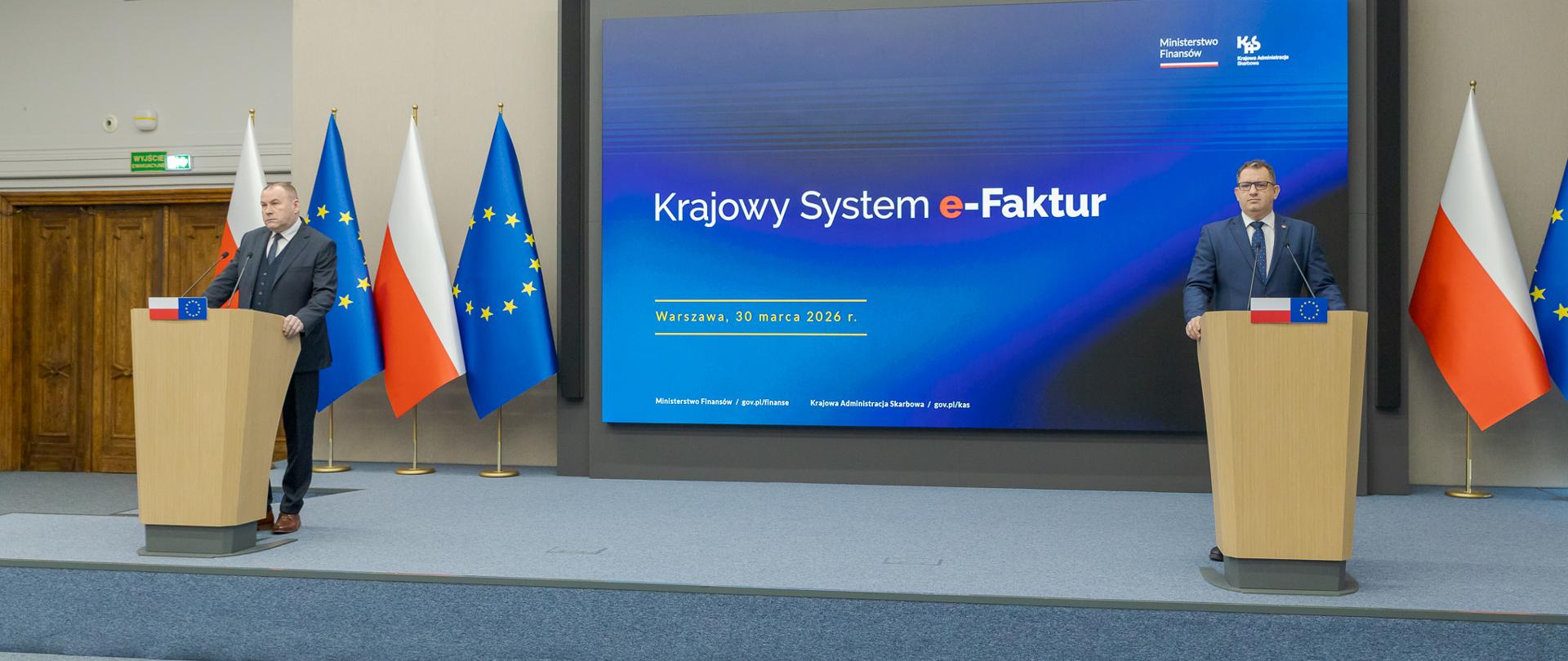 Marcin Łoboda szef Krajowej Administracji Skarbowej oraz Zbigniew Stawicki zastępca szefa KAS podczas konferencji prasowej w Ministerstwie Finansów opowiedzieli o wdrożeniu II etapu Krajowego Systemu e-Faktur przy mównicach przed dużym ekranem z napisem „Krajowy System e‑Faktur”, obok flag Polski i Unii Europejskiej.