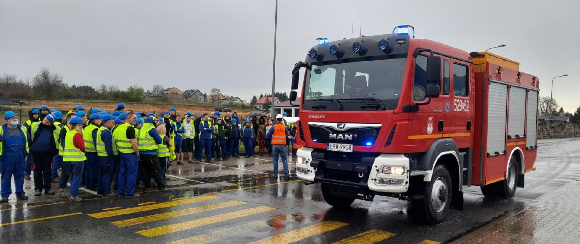Zdjęcie przedstawia grupę ludzi ubranych w niebiesko-żółte stroje robocze/magazynowe, oraz mający niebieskie kaski na głowie. Stoją oni w punkcie ewakuacji na parkingu obok nich stoi bojowy samochód strażacki.