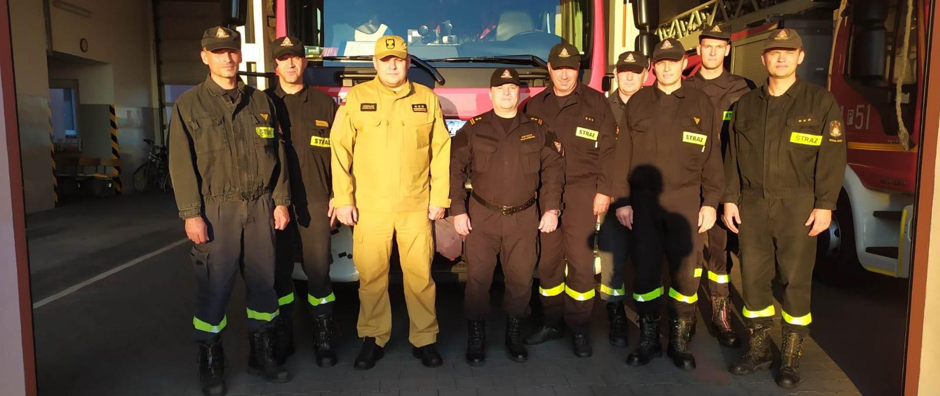 grupa strażaków w mundurach i czapeczkach dtoi przed samochodem pożarniczym