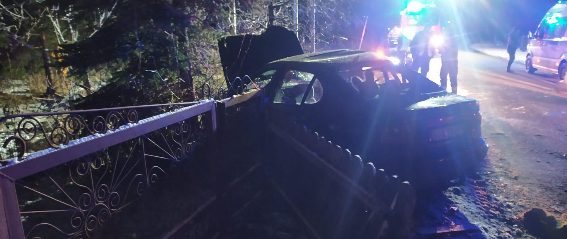 W nocy na tle niebieskich świateł stoi rozbity samochód osobowy który uderzył w ogrodzenie. W oddali samochód straży pożarnej i karetka.