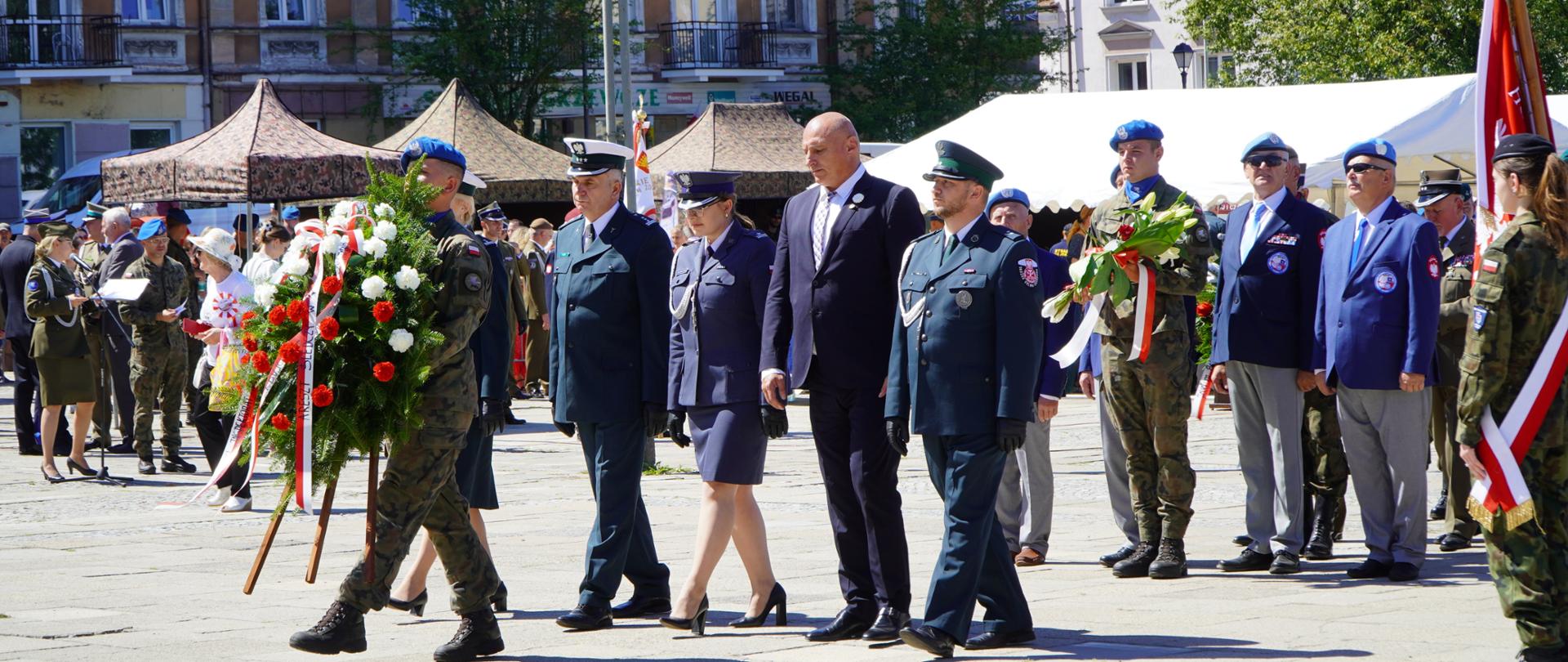 Żołnierz niesie wieniec kwiatów. Za nim delegacja sześcioosobowa.