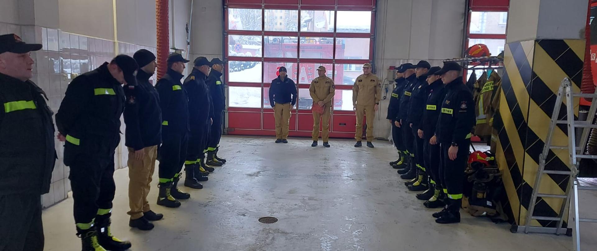 Na zdjęciu widać komendanta powiatowego PSP w Ustrzykach Dolnych, jego zastępcę, dowódcę jednostki ratowniczo-gaśniczej oraz zmianę przyjmującą i zdającą służbę. Zmiany stoją na wprost siebie a komendanci wraz z dowódcą między nimi. Zdjęcie wykonane jest w garażu JRG Ustrzyki Dolne i w tle widać bramę garażową za którą stoi samochód ratowniczo-gaśniczy. Widać również drabinę składaną, wyciąg spalin oraz wieszak na ubrania i hełmy 