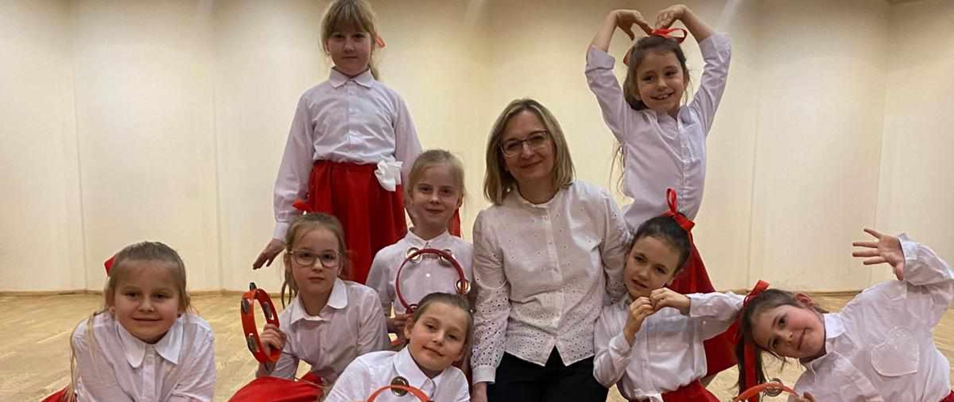 Grupa dziewcząt siedzi wokół nauczycielki na scenie