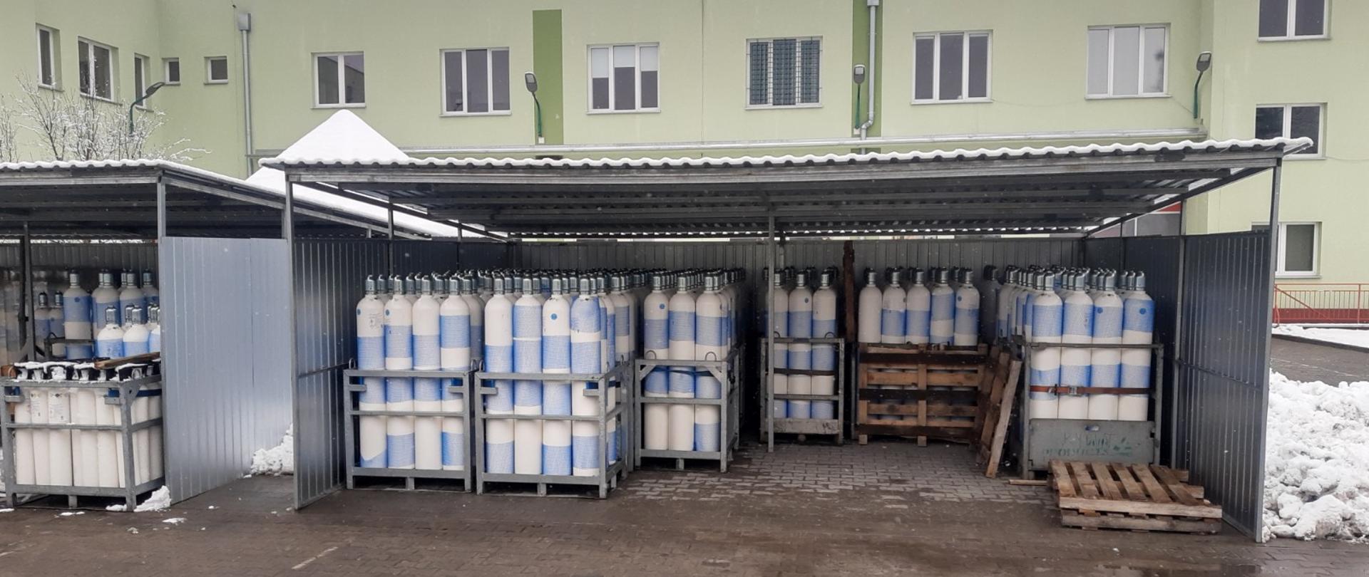 Na zdjęciu widoczny jest bank tlenu umieszczony na wewnętrznym placu JRG nr 3 Komendy Miejskiej PSP W Toruniu. Bank tworzą dwie stojące obok siebie blaszane wiaty o wymiarach 5m na 6m. We wiatach widocznych jest kilkadziesiąt stalowych koszy pełnych butli z tlenem o pojemnościach 10, 40 i 50 litrów. W tle budynek strażnicy PSP Komendy Miejskiej w Toruniu.