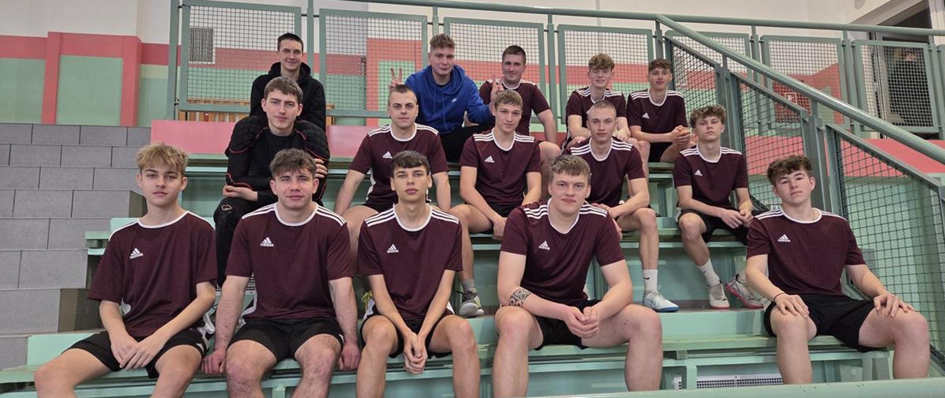 Uczniowie - członkowie szkolnej drużyny futsalu siedzą na trybunie w hali sportowej.