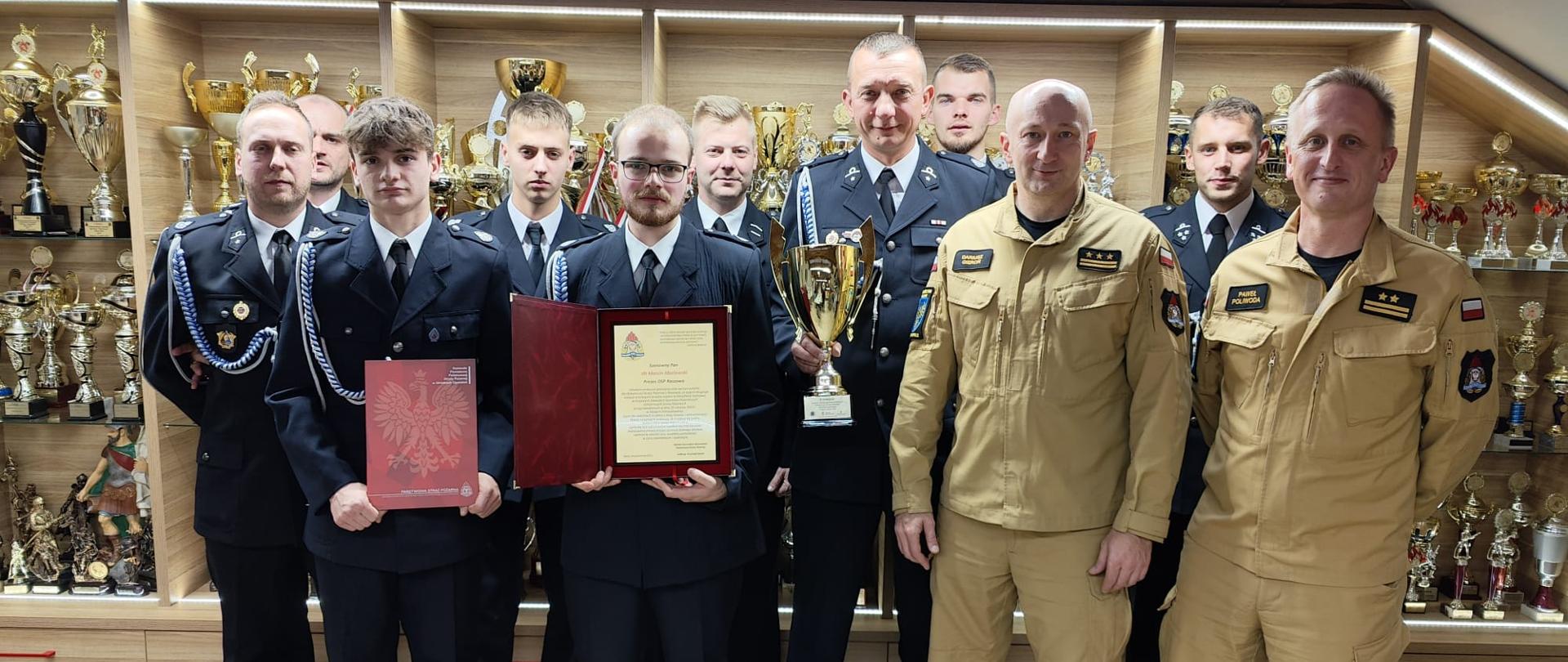 Druhowie OSP Raszowa w powiecie strzeleckim stoją w ubraniach galowych na tle wystawy pucharów wewnątrz jednostki OSP wraz z st. bryg. Dariuszem Gieroniem Zastępcą Opolskiego Komendanta Wojewódzkiego Państwowej Straży Pożarnej i bryg. Pawłem Poliwodą Komendantem Powiatowym Państwowej Straży Pożarnej w Strzelcach Opolskich. Panowie Komendanci w ubraniach służbowych koloru piaskowego. Druhowie trzymają puchar, ryngraf oraz dyplom w rękach.