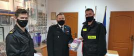 Od lewej stoi zastępca komendanta, następnie pośrodku komendant powiatowy, a od prawej druh OSP, wszyscy są ubrani w czarne umundurowanie i mają czarne maseczki na twarzy. Mężczyźni znajdują się wewnątrz komendy. Komendant wręcza druhowi czujkę dymu. Panowie zwróceni są twarzą do fotografa.