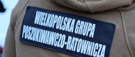 Zbliżenie na plecy beżowej bluzy polarowej z dużą czarną naszywką i białym napisem: „WIELKOPOLSKA GRUPA POSZUKIWAWCZO-RATOWNICZA”.