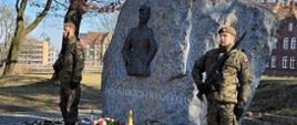 Żołnierze stoją w asyście pod pomnikiem Żołnierzy Wyklętych, pod którym leżą złożone wieńce, stoją znicze.