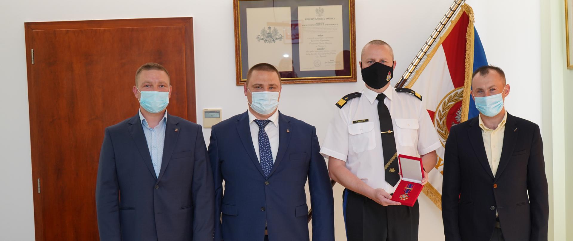 trzech mężczyzn w garniturze oraz strażak stoją przed sztandarem , strażak trzyma w ręku medal.