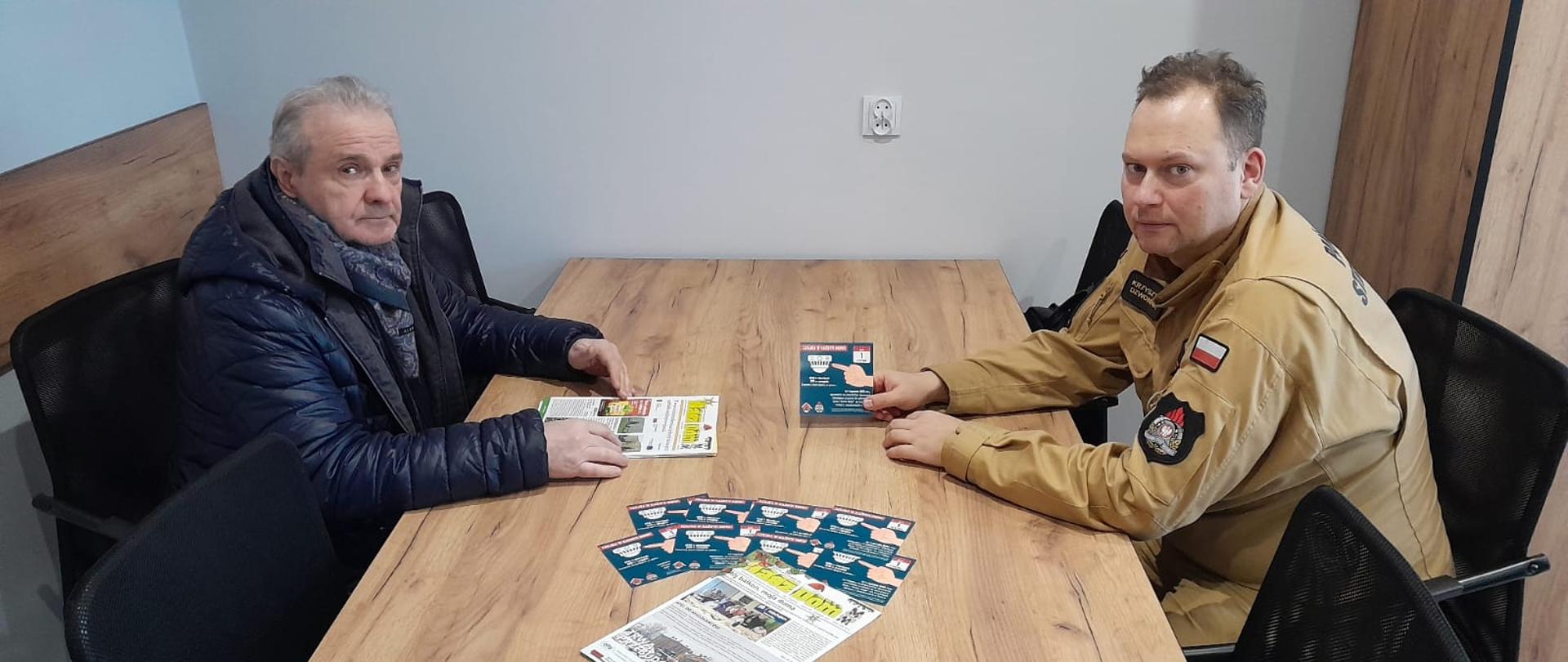 Dwóch mężczyzn siedzi przy drewnianym stole w sali konferencyjnej. Jeden z nich to strażak w umundurowaniu PSP, który prezentuje materiały informacyjne dotyczące kampanii „Czujka na straży Twojego bezpieczeństwa”. Na stole leżą ulotki i broszury związane z bezpieczeństwem pożarowym.