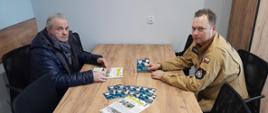 Dwóch mężczyzn siedzi przy drewnianym stole w sali konferencyjnej. Jeden z nich to strażak w umundurowaniu PSP, który prezentuje materiały informacyjne dotyczące kampanii „Czujka na straży Twojego bezpieczeństwa”. Na stole leżą ulotki i broszury związane z bezpieczeństwem pożarowym.