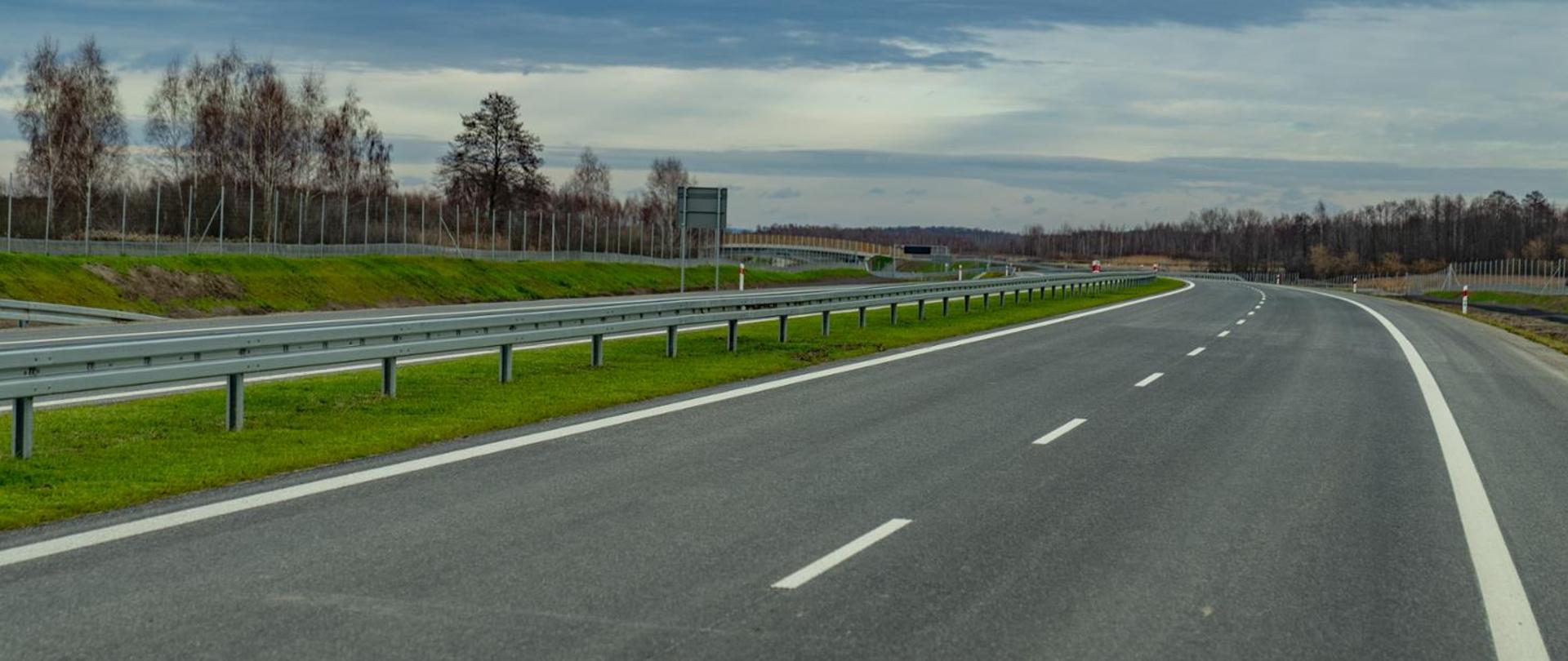 Na zdjęciu widać pustą, nową drogę ekspresową z dwoma pasami w każdym kierunku, oddzielonymi linią przerywaną. Po lewej stronie biegnie metalowa bariera ochronna i pas zielonej trawy, za którym znajduje się ogrodzenie oraz rząd drzew bez liści. Droga łagodnie skręca w prawo, a w tle widoczne są tablice informacyjne. Niebo jest częściowo zachmurzone w odcieniach szarości i błękitu.