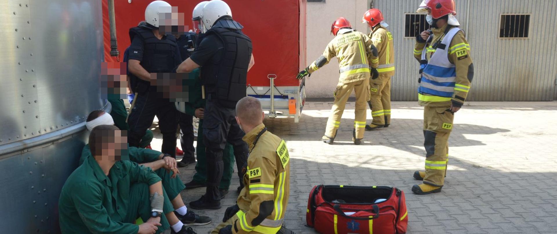 Ćwiczenia w Zakładzie Karnym w Rawiczu. Tak zwany "teren ścisły", Na pierwszym planie klęczący strażak bez nakrycia głowy, przed nim dwaj "poszkodowani", którzy zostali ewakuowani z "płonącego" budynku. Obok stoi otwarta torba PSP R1. W głębi trzech funkcjonariuszy Służby Więziennej w białych hełmach, którzy obezwładniają "agresywnego" osadzonego. Obok nich strażak - kierujący działaniem ratowniczym, a w głębi dwaj inni strażacy stojący z tyłu przyczepki koloru czerwonego.
