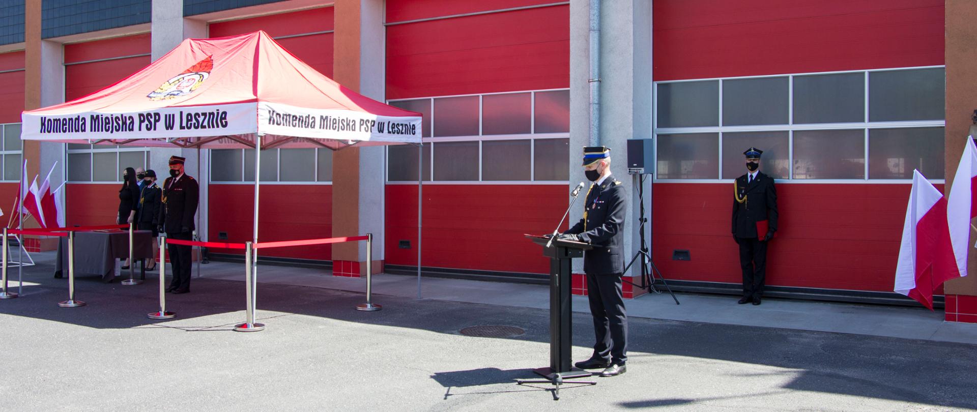 Strażak stoi przy mównicy na tle garaży komendy.