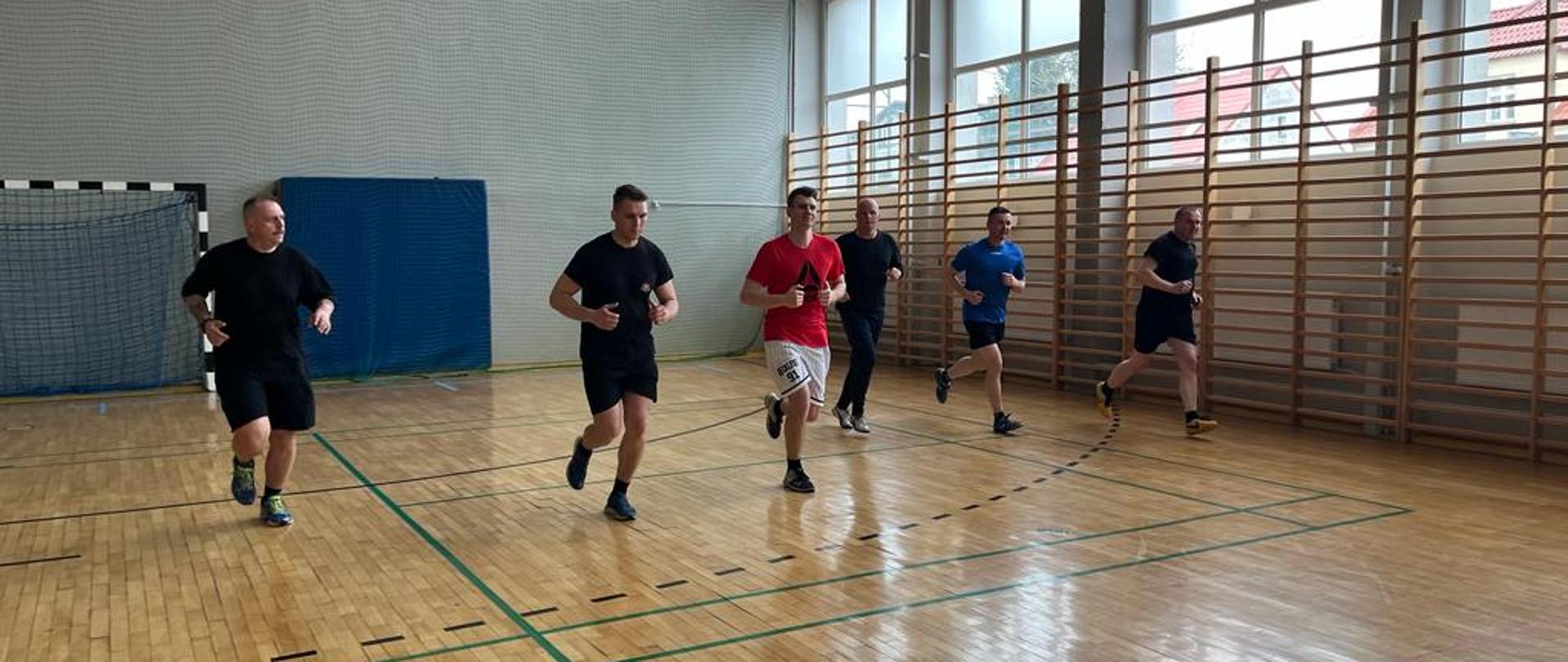 Zdjęcie przedstawia sześciu strażaków wykonujących próbę wydolnościową "beep test" na sali gimnastycznej. Na pierwszym planie funkcjonariusze zbliżają się do linii znacznikowej. Z prawej strony widoczne okna oraz drabinki gimnastyczne. W tle znajduje się bramka do piłki ręcznej, a za nią materac i siatka zabezpieczająca.