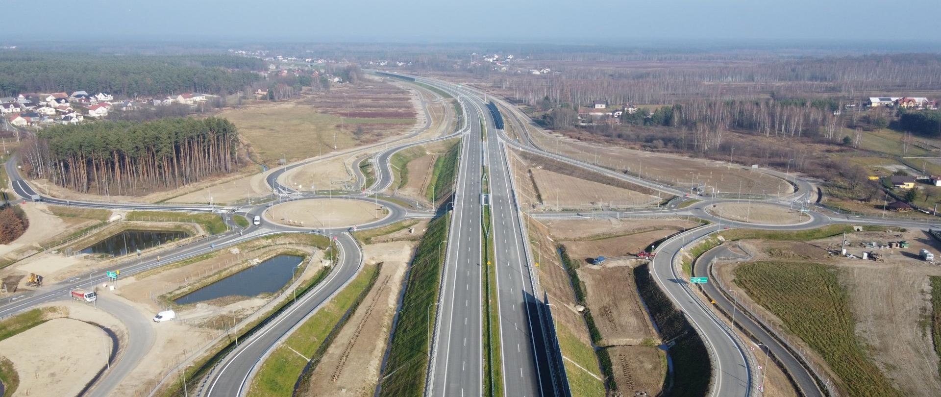 S19 węzeł Jeżowe w woj. podkarpackim, widok z powietrza na dwujezdniową drogę ekspresową w centrum kadru oraz odchodzące na lewo i prawo łącznice węzła. Po obu stronach drogi ekspresowej są duże ronda do których dochodzą łącznice węzła. 