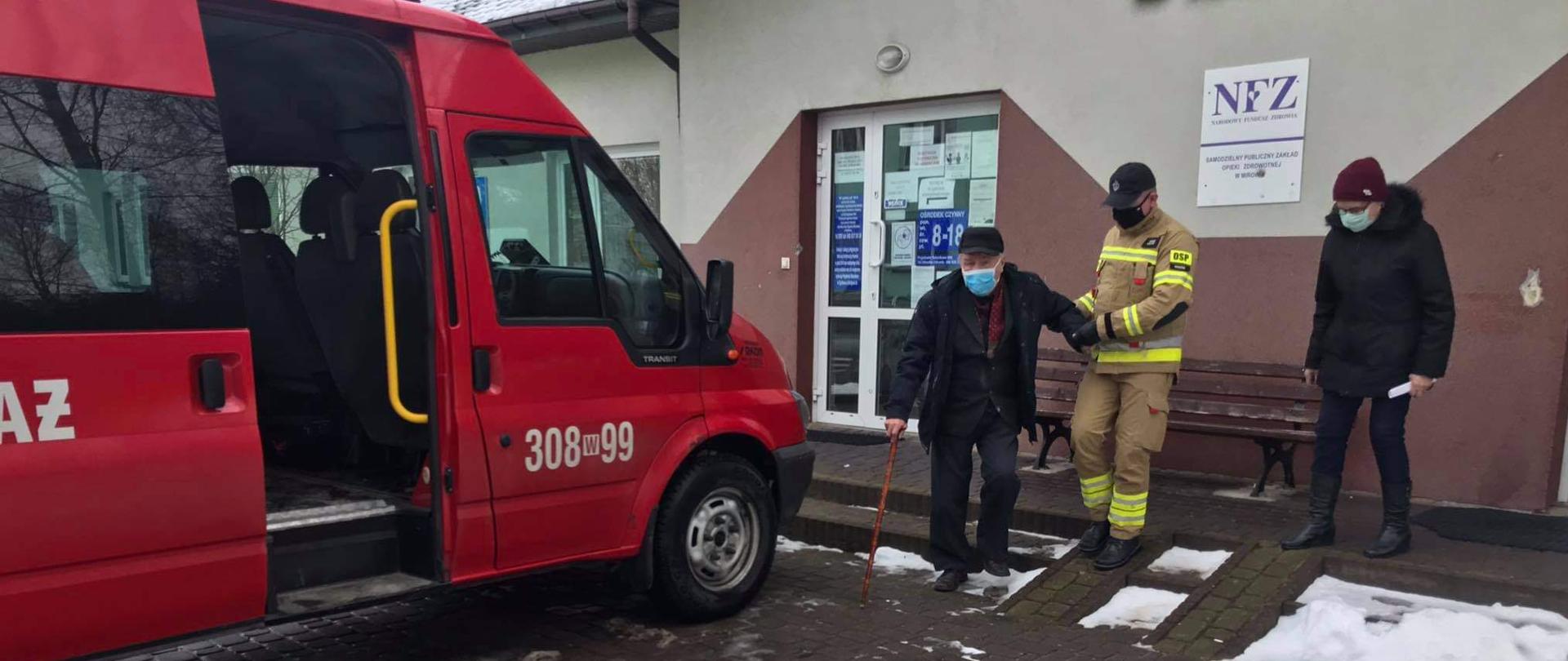 Fotografia przedstawiająca strażaka z OSP Rogów pomagającemu starszemu mężczyźnie w dojściu do samochodu strażackiego bus ze szczepienia przeciw COVID-19 celem transportu w ramach Narodowego Programu Szczepień.