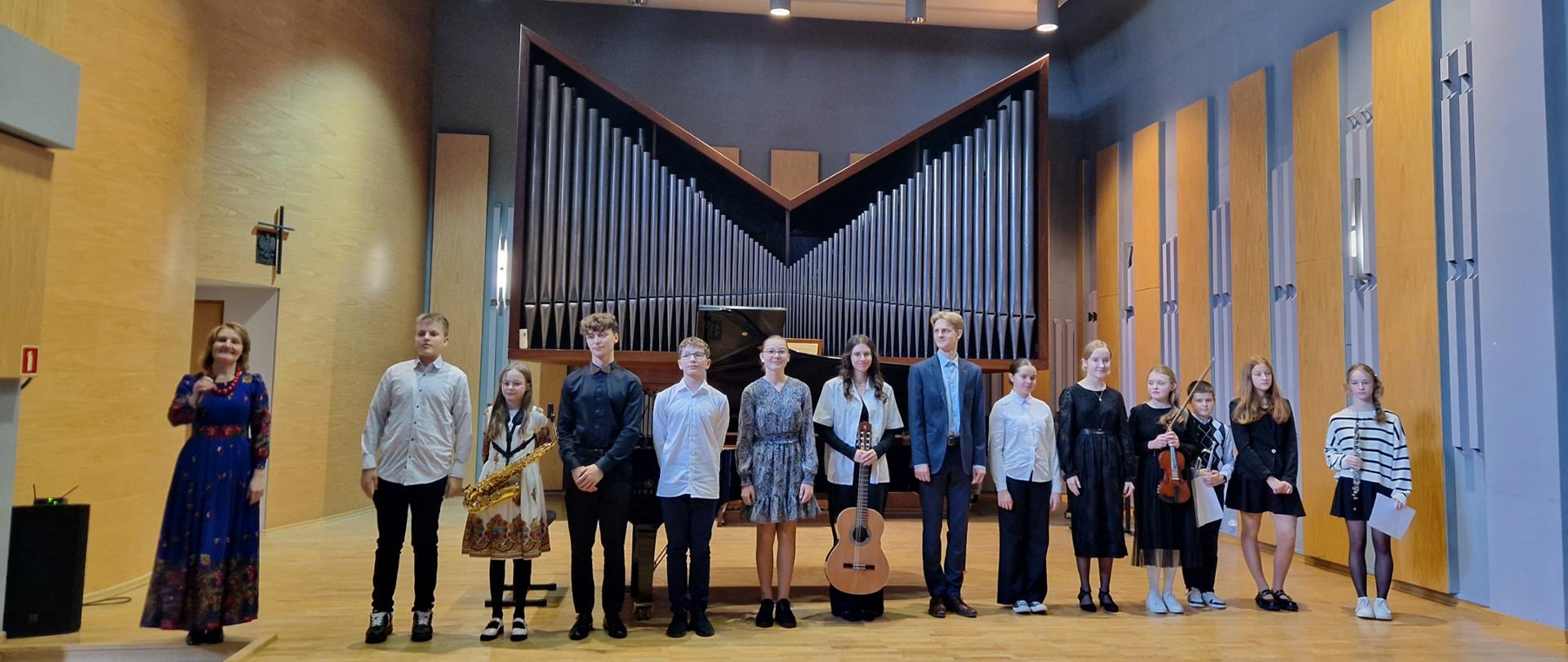Na zdjęciu widać grupę młodych muzyków stojących na scenie sali koncertowej, prawdopodobnie po występie. Oto szczegółowy opis: Stoją w równym szeregu na szerokiej, drewnianej scenie. Za nimi znajduje się ogromne tło z wielkimi piszczałkami organów – tworzą charakterystyczny, trójkątny układ i dominują całą tylną ścianę. Nad sceną wiszą prostokątne panele akustyczne oraz nowoczesne lampy skierowane w dół. Od lewej strony stoi dorosła kobieta w długiej, eleganckiej granatowej sukni w kwiaty – wygląda na prowadzącą lub nauczycielkę. Obok niej ustawiona jest grupa dzieci i nastolatków. Każde dziecko wygląda odświętnie i elegancko. Niektóre trzymają instrumenty: jedna dziewczynka trzyma saksofon, inna – gitarę klasyczną, ktoś dalej – skrzypce. Pozostali stoją z rękami opuszczonymi lub trzymają nuty czy kartki. Wszyscy patrzą w stronę publiczności, są lekko uśmiechnięci i wyglądają na zadowolonych po występie. Przed sceną widać ciemnoniebieskie fotele oraz kilka głów widzów, którzy patrzą na dzieci. Cała sala jest jasna, nowoczesna i wykonana z drewna oraz jasnych paneli akustycznych. Ogólne wrażenie: to uroczysty moment po koncercie w szkole muzycznej lub podczas młodzieżowego recitalu. Wszystko wygląda spokojnie, ciepło i profesjonalnie.