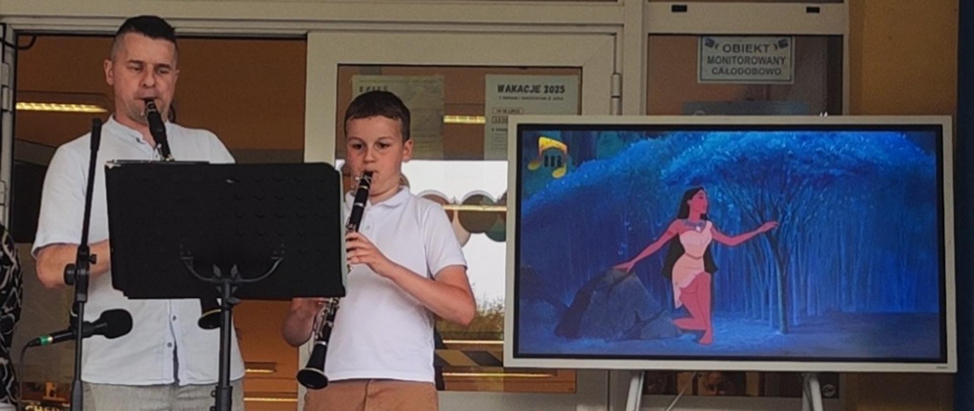 Z lewej strony grający na klarnecie mężczyzna w jasnym ubraniu. Obok niego chłopiec grający na karnecie w jasnym ubraniu. Przed nimi stoi czarny pulpit z nutami. Z prawej strony stoi ekran, na którym widać fragment bajki Pocahontas. W tle duże szklane drzwi.