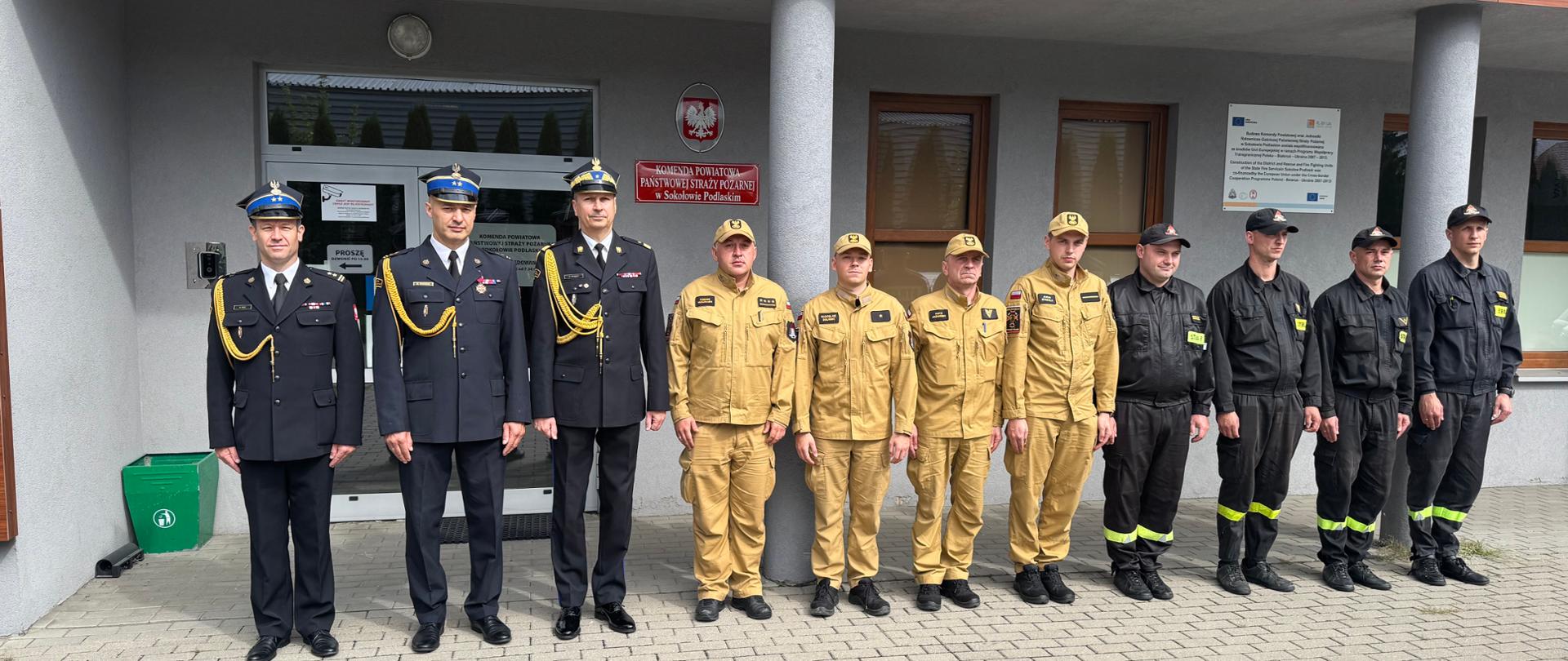 Zdjęcie przedstawia pamiątkową zbiórkę kadry i funkcjonariuszy przed wejściem głównym do budynku Komendy Powiatowej Państwowej Straży Pożarnej w Sokołowie Podlaskim (widoczna jest tablica informacyjna jednostki z godłem Polski oraz tablica informacyjna dotycząca funduszy unijnych).