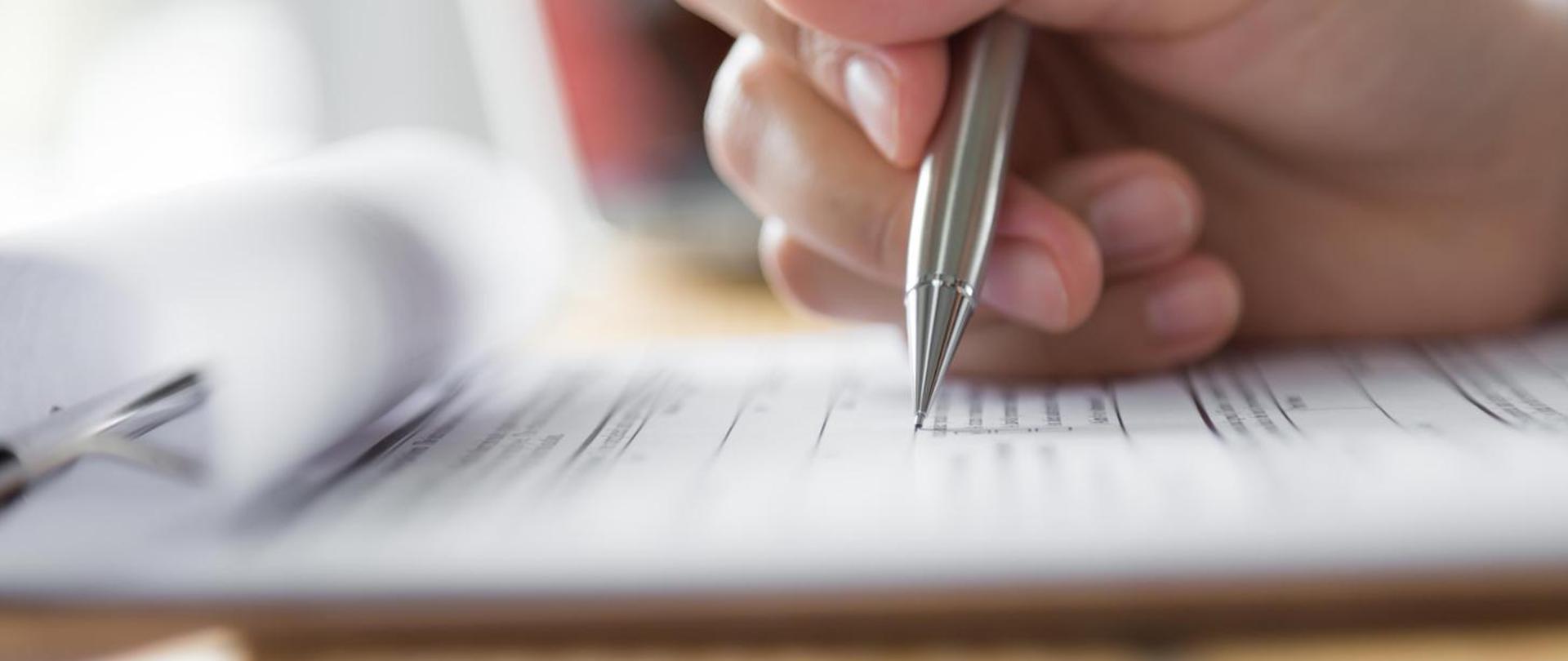 Zdjęcie przedstawia dłoń trzymającą długopis, którym podpisywane są jakieś dokumenty.