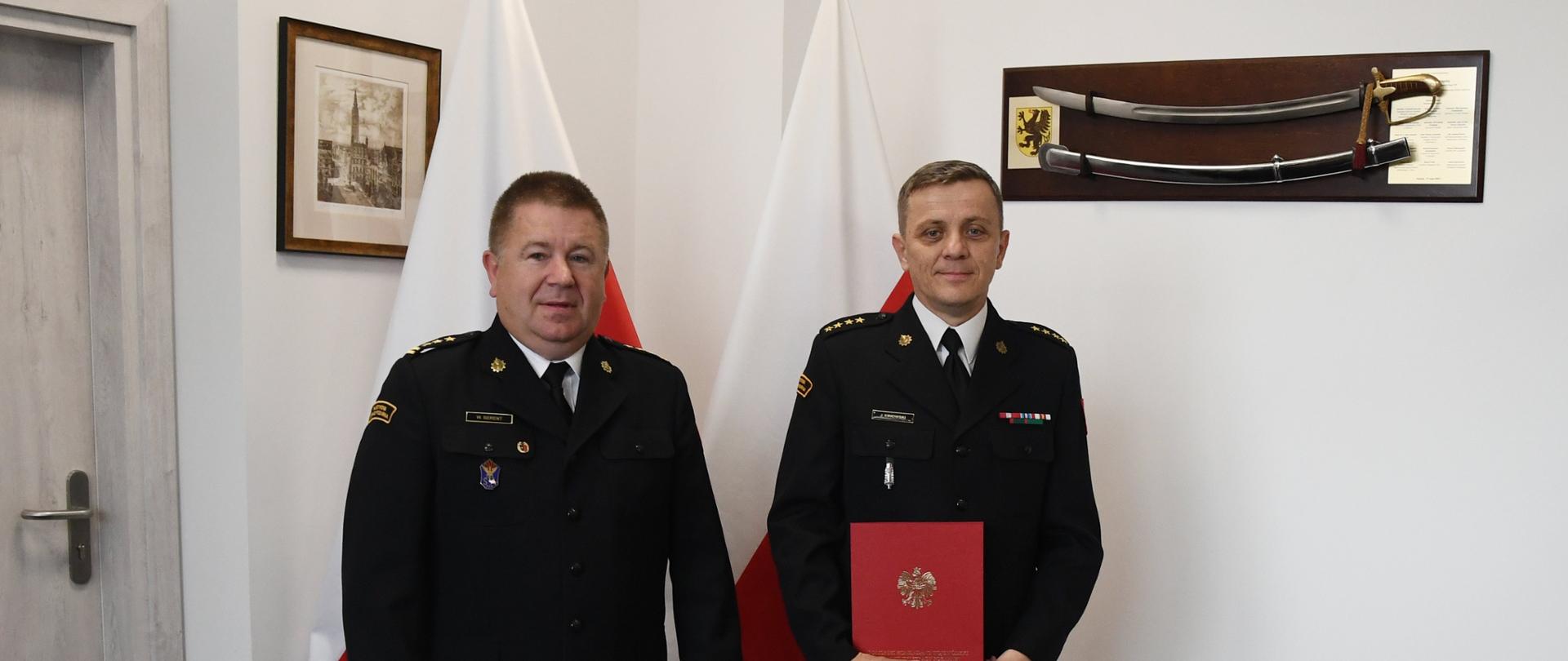 Dwóch funkcjonariuszy Państwowej Straży Pożarnej stoją obok siebie, jeden z nich trzyma czerwoną teczkę za nimi stoją dwie flagi Polski na ścianach wiszą obraz oraz szabla.