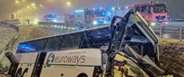 Na zdjęciu widać uszkodzony autobus, który wpadł do rowu przy autostradzie. Na pasie ruchu stoją pojazdy strażackie z włączoną sygnalizacją świetlną. Zdjęcie zrobione w porze nocnej, w trakcie opadów śniegu.