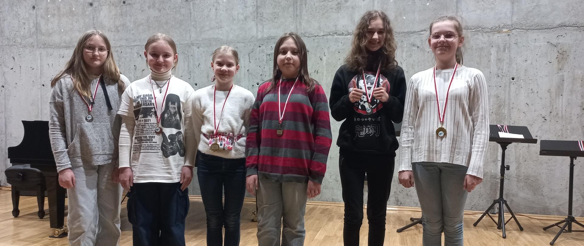 Zdjęcie przedstawia grupę uczniów stojących na scenie szkoły muzycznej. Uczennice prezentują medale otrzymane w konkursie.