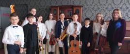 Grupa uczniów stoi zwrócona twarzą do fotografującego, w rękach trzymają instrumenty, po prawej stronie nauczycielka, w tle wzorzysta tapeta na ścianie w restauracji