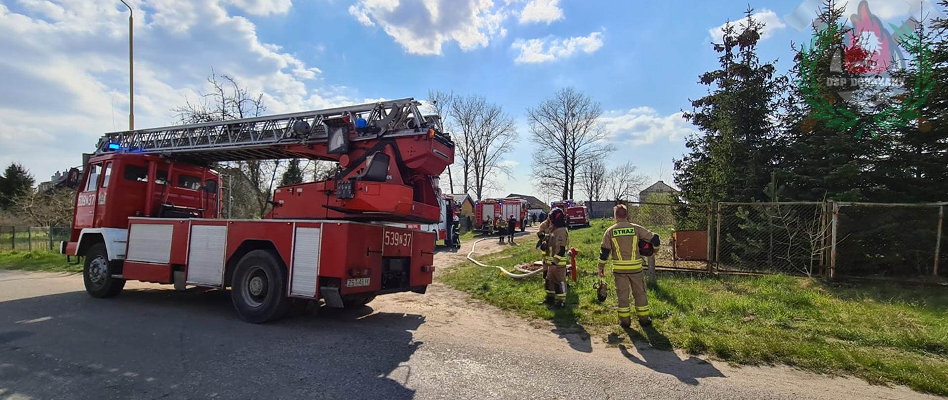 widok z drogi na pierwszym planie strażacy, samochód specjalny z drabiną
