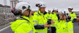 Na zdjęciu widoczna jest Ministra klimatu i środowiska Paulina Hennig-Kloska podczas wizyty studyjnej z przedstawicielami norweskiego rządu. Wszyscy ubrani są w kaski i odblaskowe kamizelki. 