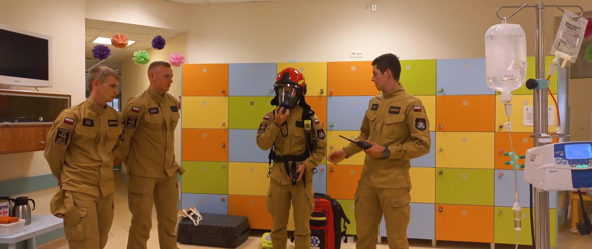 Hol Kliniki Onkologii i Hematologii Dziecięcej Uniwersyteckiego Szpitala Dziecięcego w Krakowie. Na tle urządzeń medycznych i kolorowych szafek ubrani w służbowe mundury koloru musztardowego strażacy przygotowujący pokaz i prelekcję dla chorych dzieci.