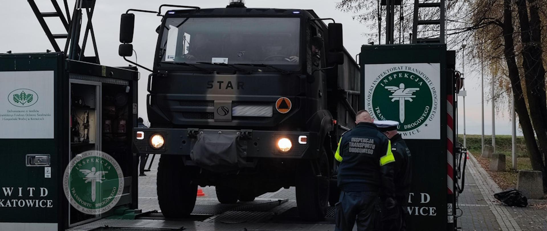 Na zdjęciu widać ciężarówkę wojskową marki STAR, stojącą na mobilnym stanowisku kontrolnym należącym do Inspekcji Transportu Drogowego (ITD). Pojazd znajduje się pomiędzy dwiema zielonymi modułowymi kabinami oznaczonymi logo ITD oraz napisem WITD Katowice.
Dwóch inspektorów, ubranych w ciemne mundury z odblaskowymi elementami i napisem Inspekcja Transportu Drogowego, wykonuje czynności kontrolne przy przedniej części pojazdu. Po lewej stronie jednej z kabin otwarte są drzwi, w których widać wnętrze wyposażenia technicznego używanego do diagnostyki.