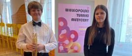 Na zdjęciu widzimy dwoje młodych ludzi stojących obok siebie w jasnym, przestronnym pomieszczeniu z drewnianą podłogą i dekoracyjnymi firankami w oknach. Chłopiec po lewej stronie ma na sobie białą koszulę, muszkę i czarne spodnie. W rękach trzyma trąbkę. Obok niego stoi dziewczyna w czarnym stroju, również trzymająca trąbkę. Za nimi znajduje się plakat informujący o wydarzeniu muzycznym. Na plakacie widnieje napis: „23. Wielkopolski Turniej Muzyczny”. Poniżej umieszczono datę wydarzenia oraz nazwę organizatora. Tło plakatu stanowi nowoczesna, abstrakcyjna grafika w odcieniach różu, czerwieni i pomarańczu, złożona z kół i miękkich kształtów.