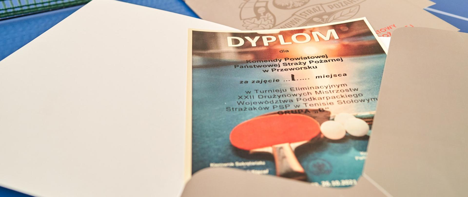 Zdjęcie przedstawia wyłaniający się z teczki dyplom uczestnictwa w turnieju eliminacyjnym tenisa stołowego.
Dyplom jest przeznaczony dla wygranej drużyny za zajęcie pierwszego miejsca.
Dyplomy oraz teczki są rozłożone na blacie stołu do tenisa.