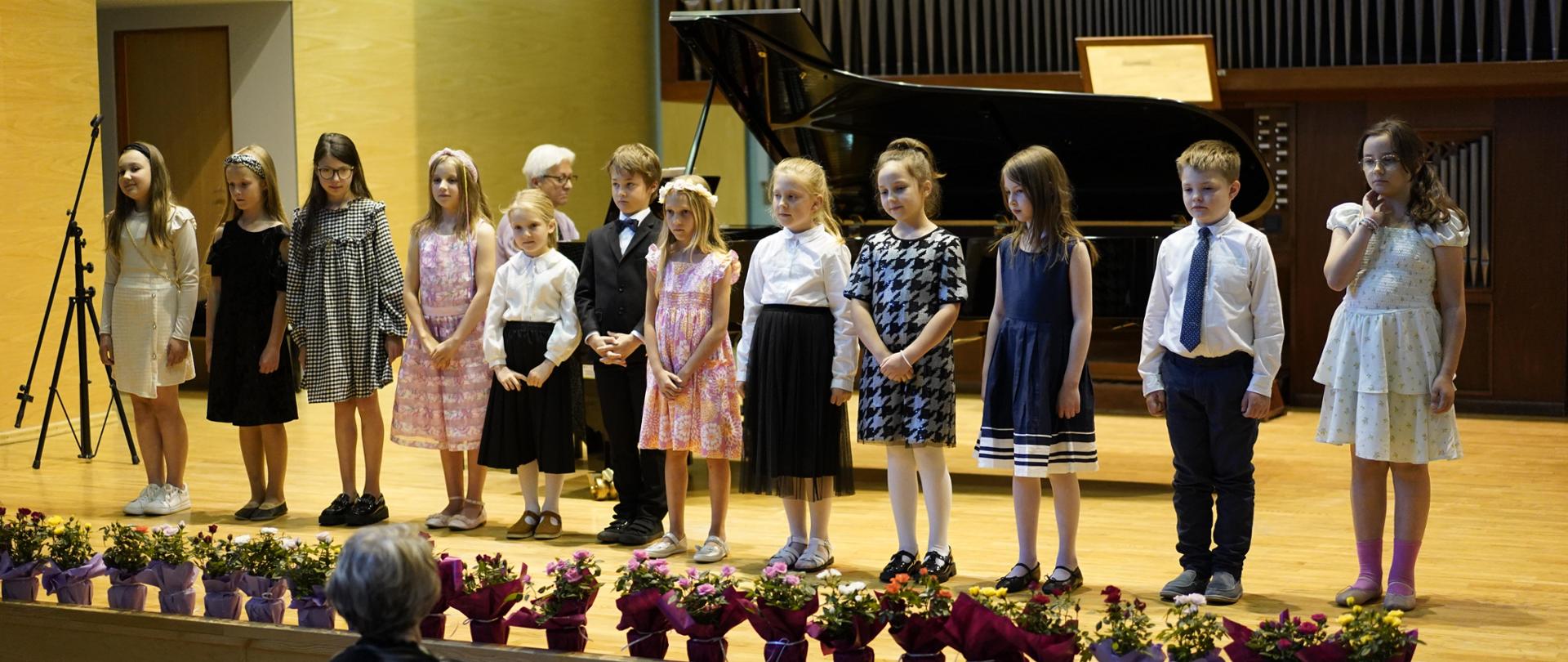 Dzieci, uczniowie klasy drugiej stoją na scenie w rzędzie. Przed dziećmi rozłożone kwiaty w doniczkach. Przed sceną część widowni. W tle za dziećmi czarny fortepian oraz organy.
