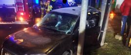 Ciemny samochód uderzył od strony kierowcy w słup, w tle widać samochód straży pozarnej