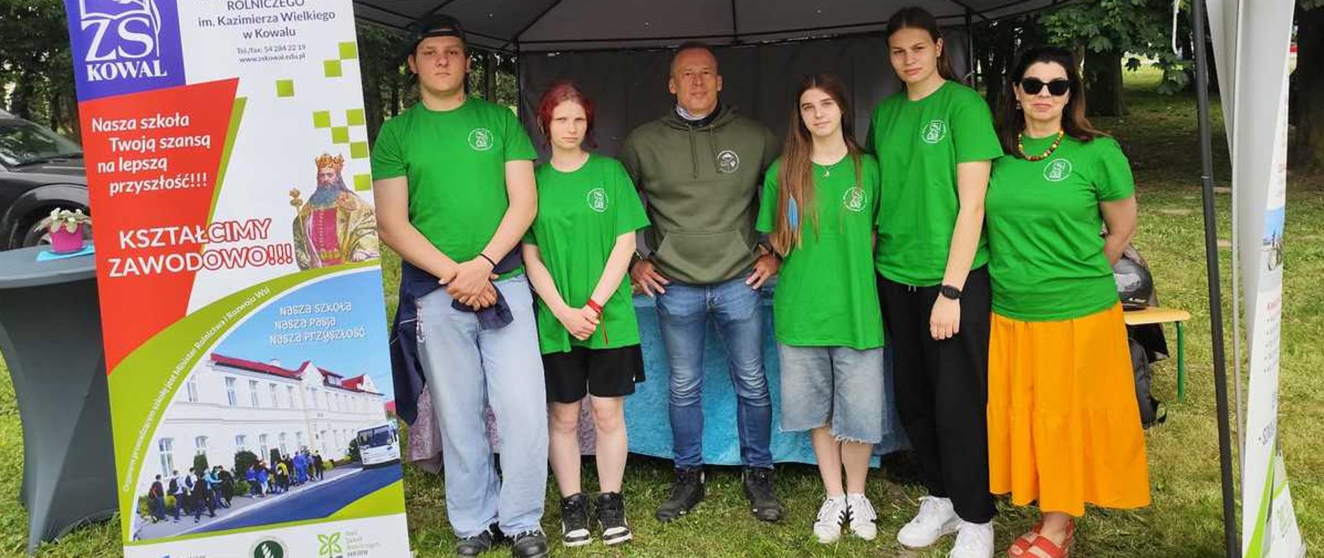 Zdjęcie przedstawia grupę sześciu osób stojących pod szarym namiotem ogrodowym (pawilonem) podczas plenerowego wydarzenia promocyjnego szkoły. Tłem dla całej sceny są gęste, zielone drzewa.Układ osób i ubiórPod namiotem, od lewej do prawej, stoją:Młody mężczyzna: ma na sobie zieloną koszulkę polo, jasnoniebieskie jeansy, ciemne buty i czapkę z daszkiem założoną tyłem do przodu. W pasie ma przewiązaną ciemną bluzę.Młoda kobieta: ma krótkie, rudawe włosy, zieloną koszulkę, czarne krótkie spodenki i sportowe buty.Mężczyzna w średnim wieku: stoi w środku grupy, ma krótkie włosy, oliwkową bluzę z kapturem, niebieskie jeansy i ciemne buty.Młoda kobieta: ma długie, ciemne włosy, zieloną koszulkę, luźne jeansowe spodenki i białe buty sportowe.Młoda kobieta: wysoka, z ciemnymi włosami spiętymi z tyłu, ubrana w zieloną koszulkę, czarne spodnie dresowe i białe buty sportowe.Kobieta w średnim wieku: ma ciemne włosy, okulary przeciwsłoneczne, zieloną koszulkę, długą pomarańczową spódnicę i czerwone sandały.Wszyscy stojący na zdjęciu (oprócz mężczyzny w środku) mają na sobie identyczne zielone koszulki z małym, okrągłym logo szkoły na lewej piersi.Elementy promocyjne i otoczenieRoll-up reklamowy (po lewej stronie)Po lewej stronie, na pierwszym planie, stoi duży, pionowy baner reklamowy (roll-up) Zespołu Szkół Centrum Kształcenia Rolniczego im. Kazimierza Wielkiego w Kowalu. Na banerze widoczne są:Fioletowe logo „ZS KOWAL”.Hasła: „Nasza szkoła Twoją szansą na lepszą przyszłość!!!” oraz „KSZTAŁCIMY ZAWODOWO!!!”.Grafika przedstawiająca króla Kazimierza Wielkiego.Zdjęcie białego, piętrowego budynku szkoły z czerwonym dachem oraz autobusu szkolnego.Otoczenie stanowiskaNamiot: szary, wsparty na metalowym stelażu, pod którym stoi stół nakryty niebieskim obrusem.Podłoże: zielona trawa z widocznymi kępkami ziemi.Po lewej stronie widać fragment szarego stolika koktajlowego oraz kawałek czarnego samochodu w tle.Po prawej stronie kadru widoczny jest fragment drugiego, białego baneru reklamowego.