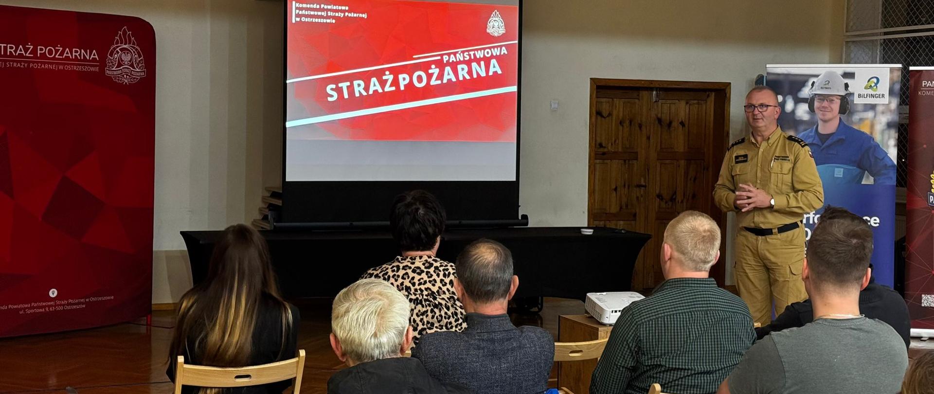 Komendant Powiatowy w trakcie wystąpienia podczas prelekcji. Na bliższym planie uczestnicy spotkania siedzą na krzesełkach. W tle banery i ekran z prezentacją. Zdjęcie wykonane w sali gimnastycznej.