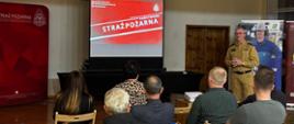 Komendant Powiatowy w trakcie wystąpienia podczas prelekcji. Na bliższym planie uczestnicy spotkania siedzą na krzesełkach. W tle banery i ekran z prezentacją. Zdjęcie wykonane w sali gimnastycznej.