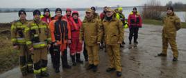 Strażacy stoją obok siebie w różnych mundurach za nimi płynie rzeka Wisła.