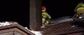 Na dachu budynku znajduje się dwóch strażaków. Przed budynkiem stoi trzech strażaków obserwujących ich pracę. 