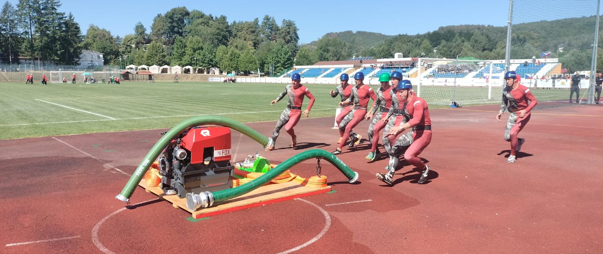 Zespół zawodników w trakcie sztafety pożarniczej. Grupa strażaków biegnie po bieżni w stronę stanowiska z motopompą, przygotowując się do podłączenia węży.