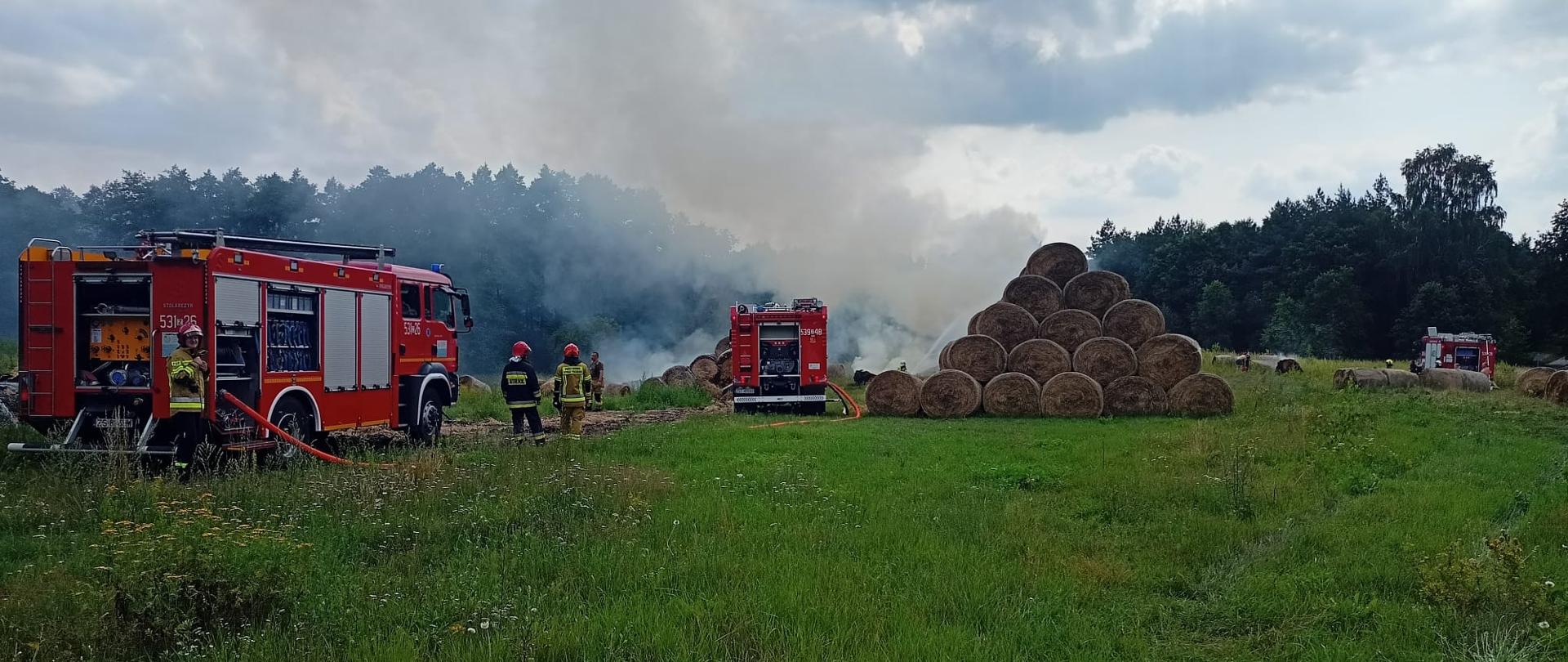 w centrum kadru sterta siana w balotach z zadymieniem, po lewo pojazdy pożarnicze oraz ratownicy