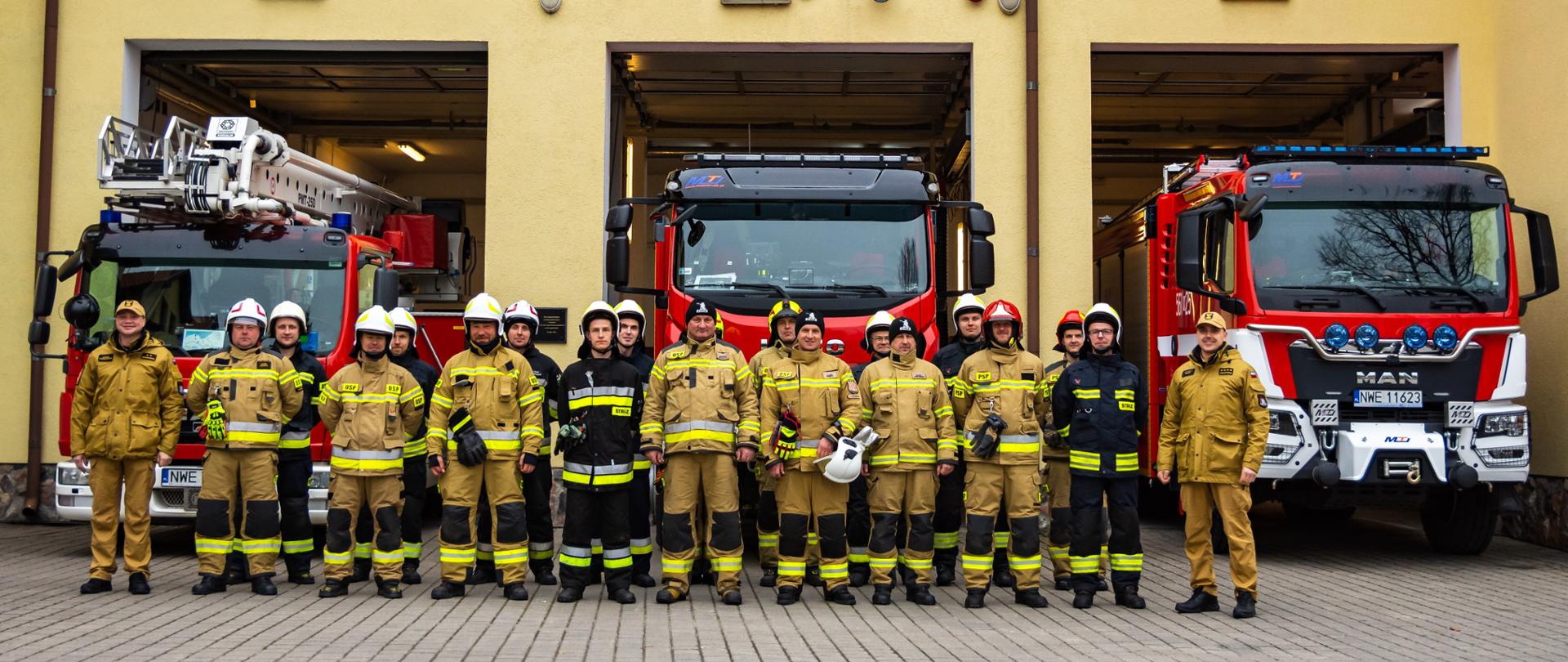 Druhowie razem z 2 funkcjonariuszami PSP stoją na placu KP tyłem do samochodów pożarniczych. 
